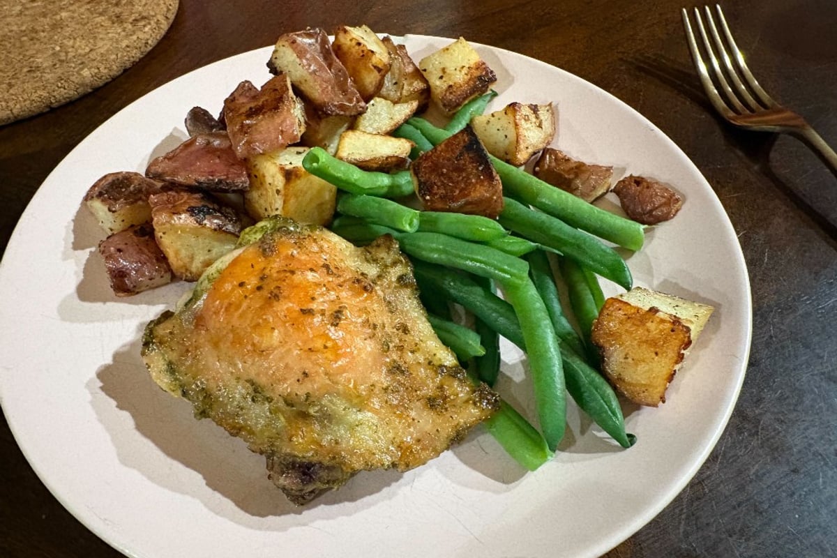 Herb roasted chicken thigh with crispy skin and green herb specks, served with crispy garlic-pepper roasted potatoes and bright green beans on a light pink plate